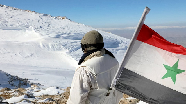 Một binh sĩ quân đội Syria với quốc kỳ Syria đang giữ vị trí tại Jabal al-Sheikh, thuộc Cao nguyên Golan.